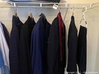Several men's jackets and pants hanging in closet, mostly dark and blue tones.