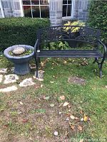 Photo of black metal garden bench with floral pattern backrest and slatted seat next to gray flower pot outside on grass.