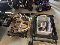 Full view of Delta Sidekick mitre saw on floor next to folding stand with plastic container holding blades on it.