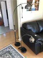 Photo showing both metal arc lamps near black leather sofa and patterned area rug, exhibiting chrome dome shades and curved arms with cords.
