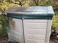 Closed view of two-door outdoor plastic storage bin with green lid and beige body, padlocked, showing outdoor wear.