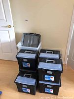 Six black portable file boxes stacked with visible handles and translucent lids, one white plastic storage bin with gray lid inside.