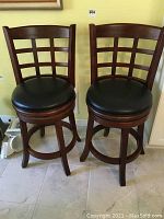 Front view of two wooden swivel bar stools with vinyl seats, lattice backs, and curved legs with circular footrests.