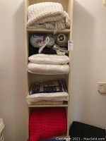 Shelves with folded textiles including cream woven afghans/blankets, storage bags, and small lamp shades.