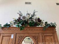Full view of the artificial flower arrangement with white and purple flowers, green ivy, placed on top of wooden carved planter.