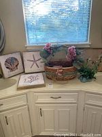 Photo of basket with pink floral accents containing towels, surrounded by framed coastal prints and a small wall plaque on cabinet countertop beneath window.