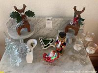 Full view of Christmas decor including wooden reindeer figurines, glass tree ornaments, ceramic Christmas tree dish, ceramic spoon, wooden nutcracker, Santa figure, and glass candle holders.