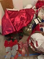 Wide view showing red satin tree skirt atop a pinecone and berry wreath, with assorted decorative bows placed nearby, and a bag of silver ornaments to the side.