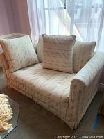 Front view of beige upholstered loveseat with three matching square pillows against window and curtains.