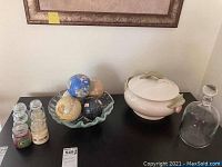 View of all items on black surface: six small jar candles, ruffled glass bowl holding five decorative globe balls, West German covered bowl with tulip handles, and clear glass decanter with stopper.