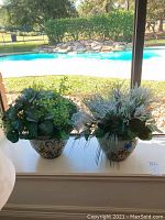 Two decorative ceramic pots with faux flower arrangements placed on a windowsill with a pool view in background.