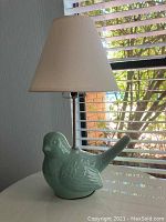 Green ceramic bird table lamp with beige lampshade placed on a white surface near a window with blind slats.