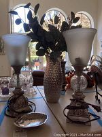 Photo showing pair of pewter look lamps with opaque frosted glass shades next to metal look textured glass vase filled with leaf flora.