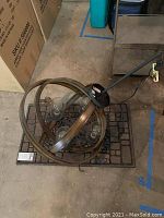 Photo showing the ceiling light fixture with four bulbs resting on the welcome mat, placed on a concrete floor with visible boxes and equipment around it.