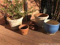 Photo showing the full set of five planters on a wooden deck with sunlight: two large plastered planters, one with a live plant, one blue ceramic planter, and two smaller terracotta pots.