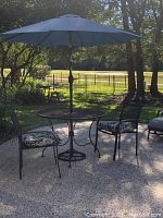 Full patio set outdoors showing round black wrought iron table with large blue umbrella and two metal mesh chairs with cushions.