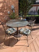 Full view of round patio table with glass top and all four cushioned metal chairs on wooden deck patio in sunlight.