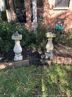 Wide shot showing two stone planters on pedestals with frog statues in garden bed area bordered by brick pavers.