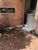 Photo of both wrought iron rocking chairs with grey cushions and the small glass-top foldable table placed on a wooden deck with brick wall background.