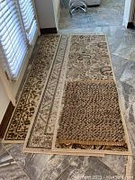 Four area rugs stacked on tiled floor near windows, two pale ivory with ornamental designs, one with braided/woven wool and jute fiber texture on top