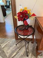 Round foldable table with metal frame and red mosaic glass top, supporting ceramic decorative boot filled with colorful artificial flowers.