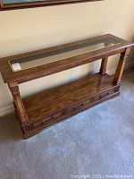 Front left angled view of wooden side table showing glass top insert, supporting columns, and bottom shelf with decorative drawer fronts.