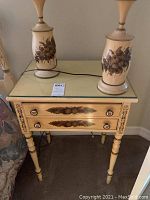 Front view of the vintage L. Hitchcock wooden bedside table with a glass top, two drawers featuring floral decorative accents, and two lamps resting on top.
