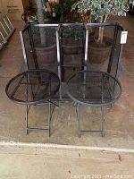 Two black metal round folding tables with mesh tops and folding legs placed side by side.