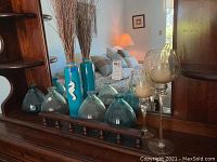 Wide shot displaying four recycled glass decanters in blue and green hues, light blue seahorse vase, and two clear glass candle holders visible on wooden shelf