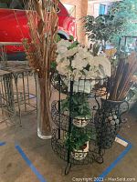 Photo of vases including two tall clear glass vases and one large dark perforated metal vase with tall faux reeds along with black three-tier wire shelf with faux plants.