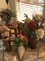 Photo showing metal and glass vases with various faux flowers and a wooden stool near window blinds.
