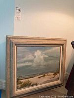 Framed beach scene painting on canvas showing sand dunes, grasses, cloudy sky, rustic wooden frame.