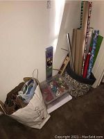 Photo of gift wrapping supplies arranged by wall including rolls of assorted wrapping paper in decorative fabric storage bin, a plastic box of bows, and a white bag containing gift bags.