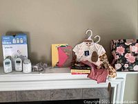 Wide view of all items on mantle: VTech baby monitor units, cords, baby clothes on hangers, books, greeting cards, and a floral box