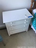 White chest of drawers with three drawers and two white knobs per drawer shown from front, on carpet.