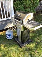Full view of black and stainless steel KitchenAid gas grill including side shelves and propane tank next to grill on grass.