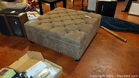 Square brown upholstered ottoman shown from a side angle, highlighting tufted top and nailhead trim detail, placed on a reddish-brown floor.