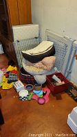 Photo shows metal pet crate disassembled with white padded base leaning against a wooden cabinet, assorted pet beds and mats piled nearby along with ceramic bowls and pet toys including a pink toy and a chew toy.