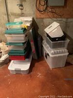 Stack of various sized plastic storage bins with locking lids and different colored lids including white, green, teal, and red. Some bins are stacked, placed on the ground in basement setting.