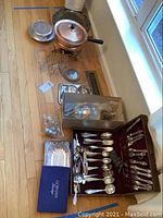 Overall view of flatware sets, chafing dish, kitchen stands and boxed Graham flatware on hardwood floor by window.