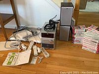 Photo of Nintendo Wii console and accessories on floor including remotes, balance board, and JVC audio system with speakers, plus VHS tape boxes