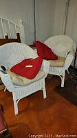 Pair of white wicker chairs with beige seat cushions and red back cushions. Chairs placed against basement wall, showing paint chipping on legs and arms.
