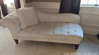 Full view of beige upholstered chaise lounge with tufted button seat and attached cushion, sitting on light carpet in front of windows.
