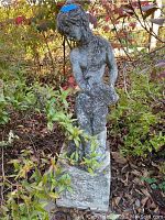 Full view of the weathered cement female garden statue surrounded by garden foliage.