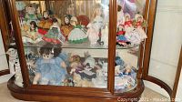 Overview of two lower shelves in curio cabinet displaying multiple dolls including Madame Alexander dolls mostly in traditional dresses and some unnamed china dolls and Effanbee dolls.