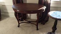 Side view of dark wood oval table with carved legs and curved supports, placed on carpet near curtains.