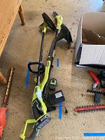 Full view of the Ryobi string trimmer and limb saw laid on floor with battery and charger nearby.