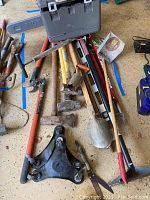 Wide view of lot including Ryobi chainsaw case, limb clippers, sledge hammers, shovel, tamper and tool with wheels laid on floor