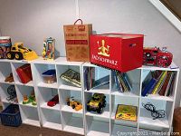 Wide shot showing white cubical shelving unit holding various toys including toy vehicles, books, and games along with FAO Schwarz toy box on top.