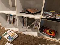Shelved DVDs and Wii/Wii U/GameCube game cases in white shelving cubbies on carpeted floor.